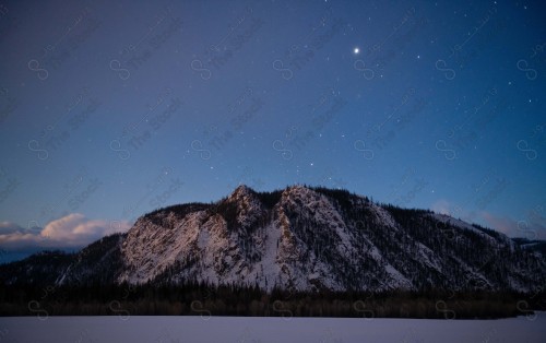 A mountain topped by a group of trees while the stars appear shining in the night sky, a mountain is covered with snow while surrounded by a group of trees