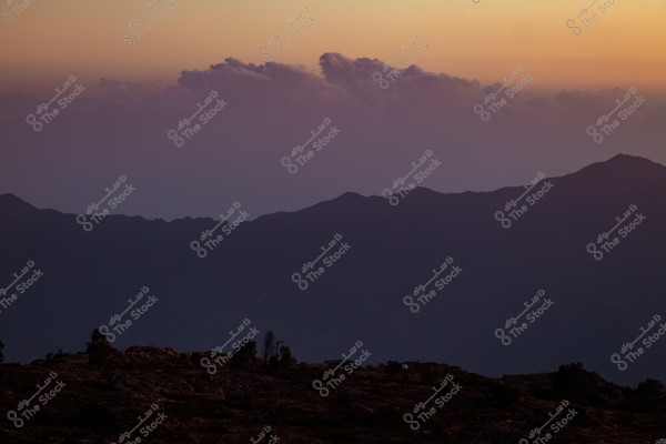 Image of a natural landscape featuring mountains with dark silhouettes in the foreground, and a pale sky adorned with gradients of orange and blue along the horizon, indicating either sunset or sunrise. Clouds line the horizon, adding a dramatic touch to the scene.