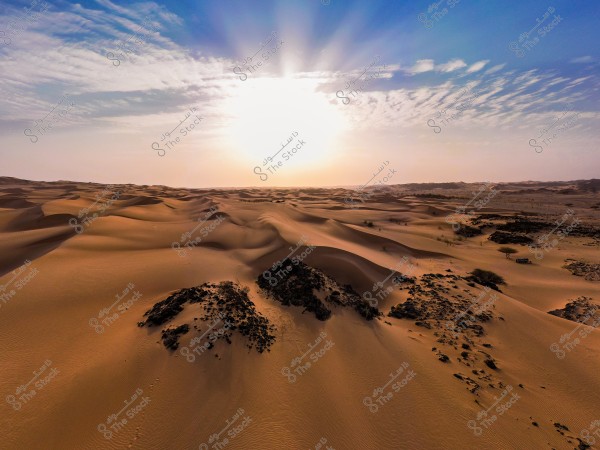 A vast desert landscape with golden sand dunes stretching in all directions under a blue sky with scattered clouds. The bright sunlight casts a warm glow over the sands, with scattered desert vegetation and black rocks visible in the distance.