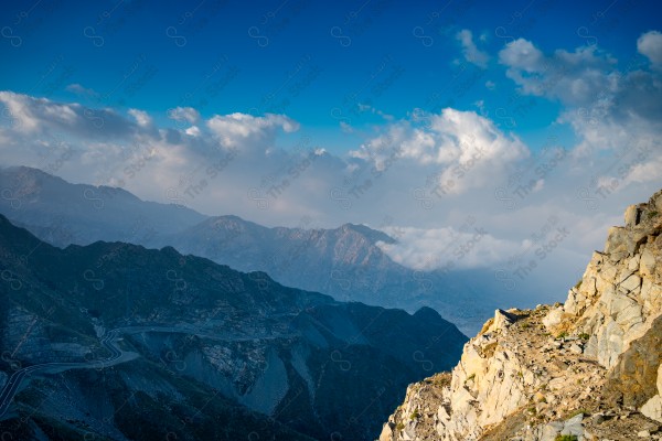 A snapshot of a series of towering rocky mountains and paved roads extending in the Al Hada Mountains in Taif, showing a cloudy sky during the day, mountain heights, Taif Mountains, mountain range