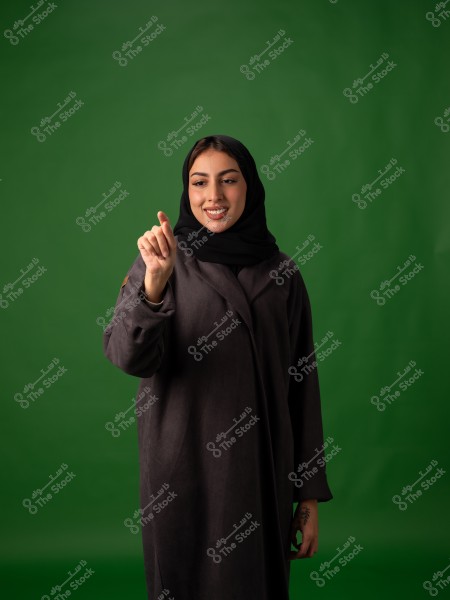 امرأة ترتدي حجاباً تشير بإصبعها وهي مبتسمة, مع خلفية خضراء.