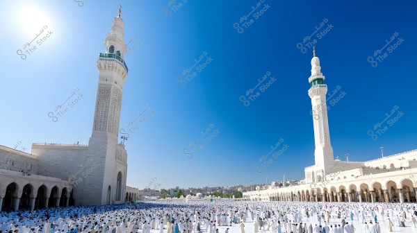 Image of the courtyard of the Prophet\'s Mosque in Medina, Saudi Arabia, with a large number of people wearing traditional white clothing, symbolizing the attire for Ihram during Umrah or Hajj rituals. Two tall minarets with Islamic architectural design are visible, set against a clear blue sky.