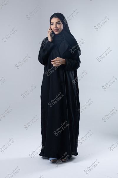 An image of a woman wearing a black abaya and black hijab, standing in a studio with a gray background, talking on a mobile phone. She appears confident and gently smiling, conveying a sense of social interaction or conversation.