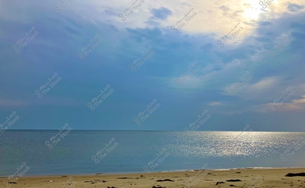 A serene beach scene in the evening with cloudy skies and calm sea waters. The sun is faintly visible behind the clouds, casting a golden glow on the water's surface. The sandy beach is in the foreground with some scattered seaweed.