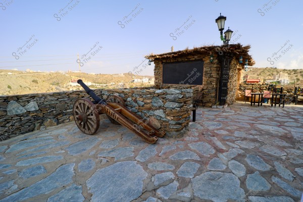 Image depicting an old cannon with wooden wheels placed on a stone pavement. The cannon is situated next to a low stone wall enclosing a small courtyard with a small stone building and a roof covered with branches. There are a few tables and chairs in the courtyard and traditional lanterns on poles. The background shows hills and small scattered buildings on the horizon with a clear blue sky.