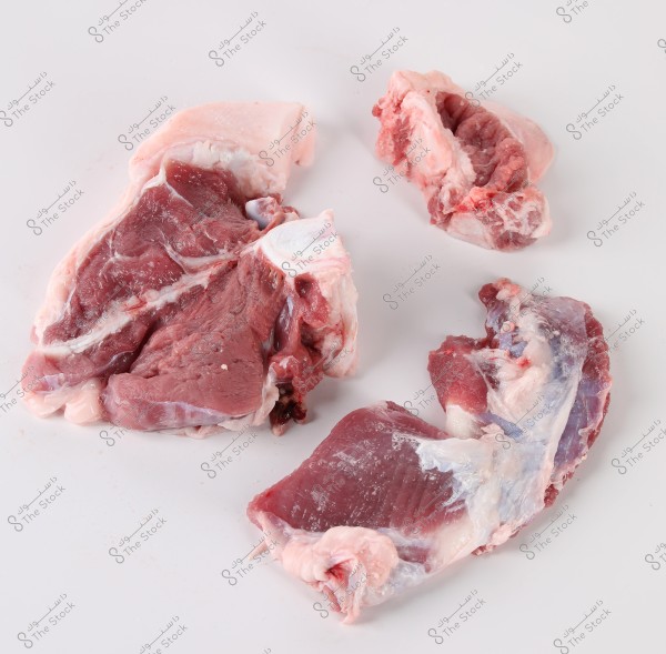 Three pieces of raw meat placed on a white surface. The meat pieces have some fat and bone. The meat appears dark red with white fatty portions.