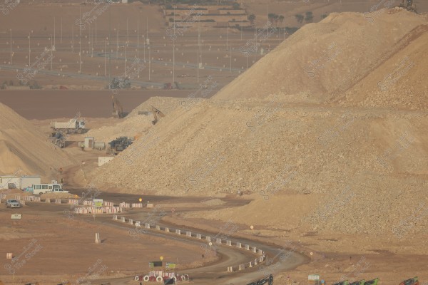 The image depicts a construction site with large mounds of dirt and rocks, along with excavators and trucks in the background. There are signs and traffic signals around the area, and a winding road runs through the site. The ground is covered with dry, brown soil.