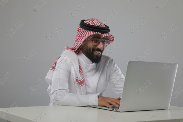 A man wearing traditional Gulf attire, with a white thobe, red keffiyeh, and black agal, sitting at a laptop on a white table. He is smiling and appears happy while using the computer. The background is gray and calm.