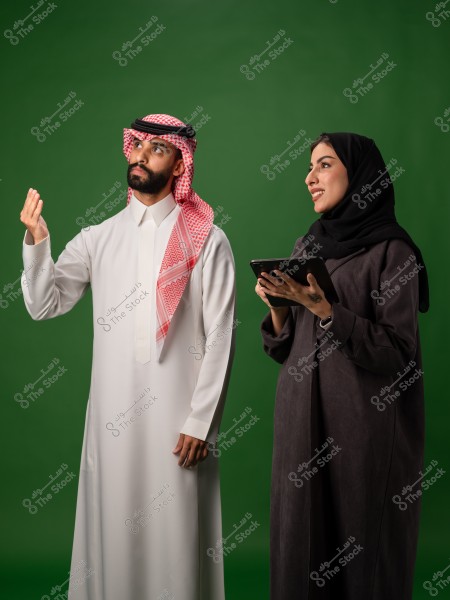 رجل وامرأة يرتديان الزي التقليدي يقفان مع خلفية خضراء, المرأة تحمل جهازًا لوحيًا.