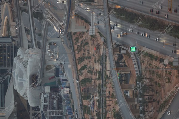 صورة جوية لبنية تحتية حضرية معقدة في مدينة كبيرة, تتضمن مجموعة من الطرق السريعة المليئة بالسيارات وجسور موجهة بتصاميم معمارية مستقبلية. تظهر على اليسار بنية معمارية بيضاء كبيرة بسطح منحني يشبه زهرة اللوتس. هناك أشجار نخيل متناثرة ومناطق خضراء على طول الطرق. يبدو أن الموقع يكون حيويًا مع حركة مرور نشطة, مما يعطي انطباعًا لمدينة متطورة.