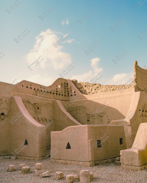 Image of an ancient mud-brick structure featuring traditional geometric designs with triangular openings. The building is located in a desert area under a clear blue sky with a few clouds. There are small stones scattered on the ground.