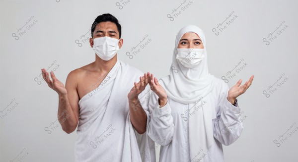 A photo of a man and a woman standing together in traditional ihram clothing. The man is wearing a white wrap without a shirt, and the woman is wearing a white headscarf and abaya. Both are wearing white face masks and appear to be in a prayer position, gently raising their hands. The background is simple and uniform, highlighting the details of their traditional attire.