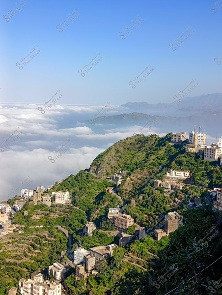 A natural landscape of a mountain covered with greenery, with houses integrated into its slopes. Thick white clouds are visible on the horizon in the background. The sky is clear and blue above, creating a serene atmosphere.