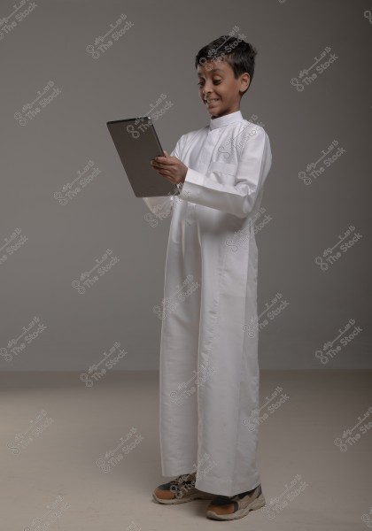 An image of a young boy wearing a traditional white thobe, standing with a smile on his face while holding a tablet in his hands. He appears to be enjoying interacting with the device. The background is simple and gray.
