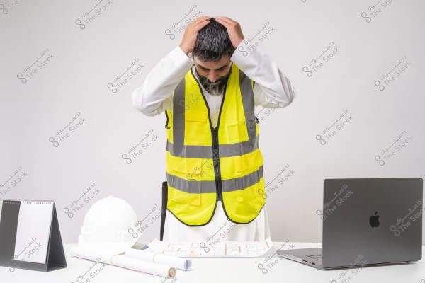 An engineer wearing a yellow reflective vest stands in front of a white desk, placing his hands on his head. In front of him are a white hard hat, engineering blueprints, and a laptop. The engineer is wearing a white robe, suggesting the image may be from an Arab country.