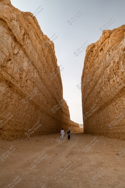 The image shows a large rocky crevice resembling a canyon with high sandy rock walls, and a sandy path in the center. In the background, two people are walking along the path; one is wearing a traditional Saudi white thobe, and the other appears to be wearing a black abaya. The sky above is clear and blue.