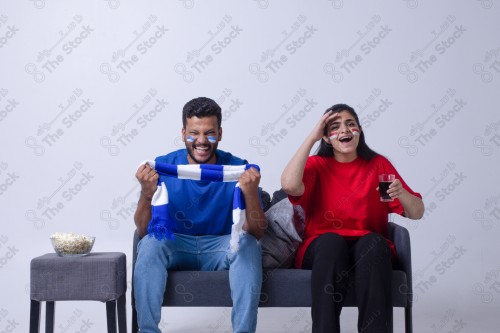 Portrait of a Saudi young man and woman watching a match and cheering their favorite teams cheerfully while having a snack on a blue background, the World Cup.