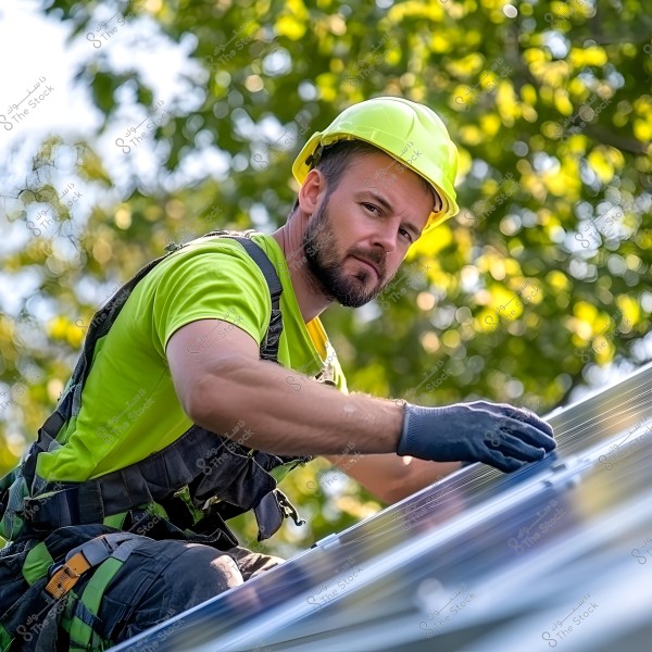 The image shows a man wearing a yellow safety helmet and a light green shirt. He is working on installing or maintaining solar panels, indicating a focus on renewable energy installation. He is also wearing gloves for protection while working. The background features green trees under sunlight.