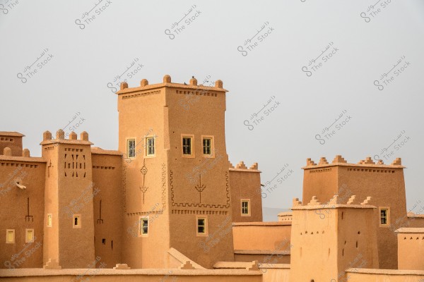 Image of a traditional fortress made of light brown mud bricks, featuring small windows and simple decorations on the walls. The architectural design showcases traditional patterns, reflecting the style of ancient Arab fortresses in desert regions. The sky behind the buildings is gray, indicating cloudy weather.