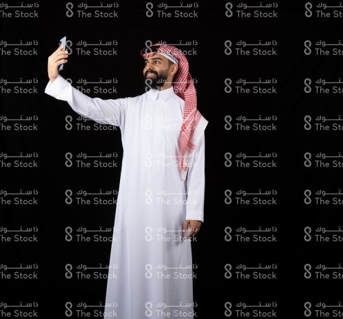 ربورتريه جل سعودي  يقف على خلفية سوداء يرتدي الثوب يحمل جوال بيديه ويلتقط لنفسه صورة وهو مبتسم ،
رجل يلتقط لنفسه صورة بكميرا الجوال الامامية