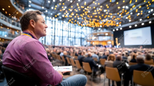 The image shows a man sitting in a large conference hall filled with attendees. The man is wearing a purple shirt and has a lanyard. The hall is decorated with small lights on the ceiling, and there is a large screen in the background displaying a blurry presentation. The audience is seated in organized rows of chairs in the hall, which features large glass windows.