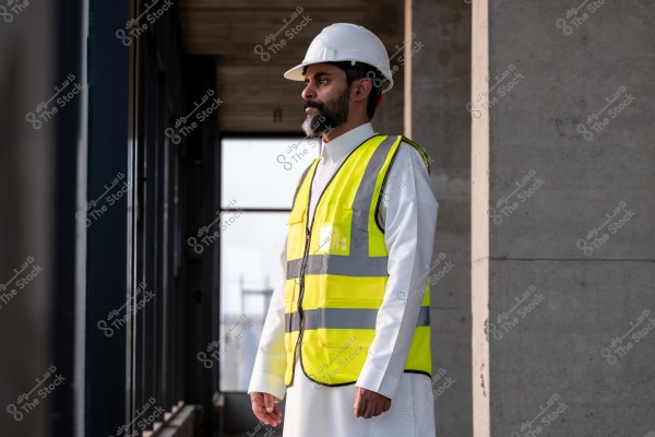 An image of a man standing inside a building under construction. He is wearing a white hard hat and a yellow reflective safety vest over a white thobe, suggesting he works in construction or engineering. The interior details show concrete walls and large windows, giving the impression of a construction site environment.