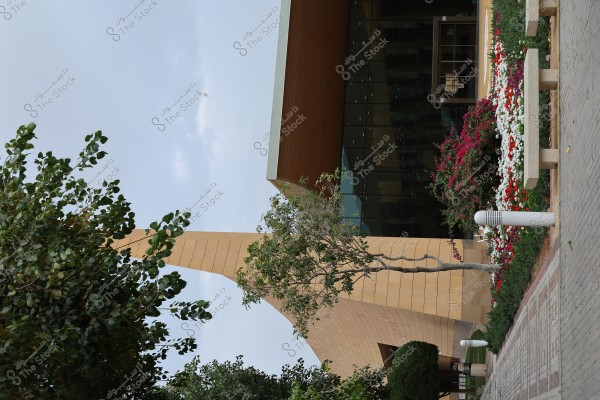 A modern building with a distinctive architectural design on a cloudy day. The building features a large glass facade and a wide entrance. The front gardens are adorned with a variety of colorful flowers, including white, red, and pink blooms. Trees are present on the sides, with brick paver walkways near the entrance.