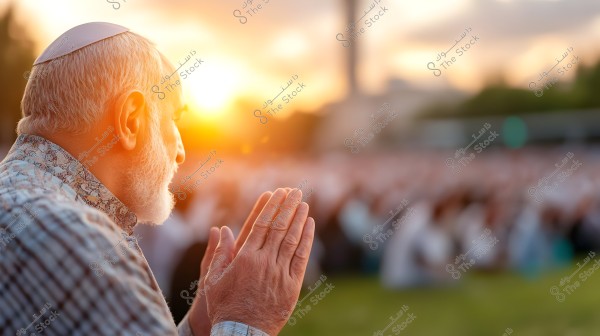 An elderly man with a white beard wearing a small cap and a patterned shirt is raising his hands in prayer outdoors at sunset. The background is blurred, showing a large gathering of people seated on the grass.