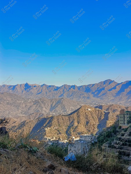 مشهد طبيعي يظهر سلسلة جبال تحت سماء زرقاء صافية. في المقدمة, توجد منطقة سكنية مبنية على المنحدرات الجبلية, مع انتشار النباتات البرية والأعشاب. الجبال تمتد في الخلفية, مع وجود بعض الظلال التي تلقيها أشعة الشمس.