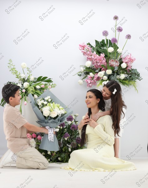 The image shows three people in a lovely floral setting. A woman dressed in a pale yellow dress is sitting with a girl behind her in a short white dress. In front of them, a young boy is presenting a bouquet of white flowers. The background features various floral arrangements of pink, purple, and white flowers, adding a touch of beauty and nature.