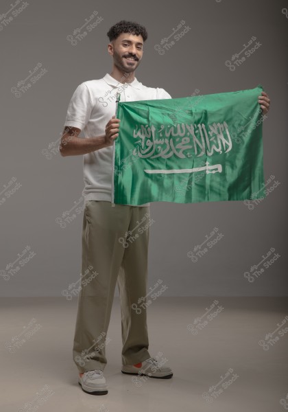 An image of a man standing and smiling while holding the Saudi Arabian flag, which is green and inscribed with the words \"There is no god but Allah; Muhammad is the messenger of Allah\" with a white sword below. The man is wearing a white shirt and khaki pants with sneakers. The background is neutral and smooth.