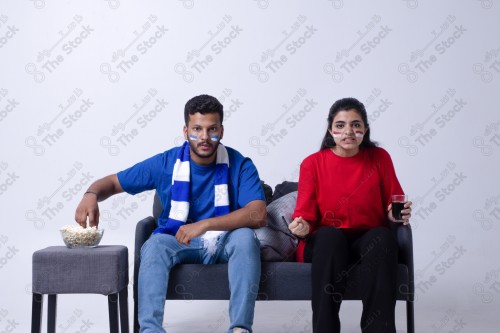 Portrait of a Saudi young man and woman watching a match and cheering their favorite teams cheerfully while having a snack on a blue background, the World Cup.