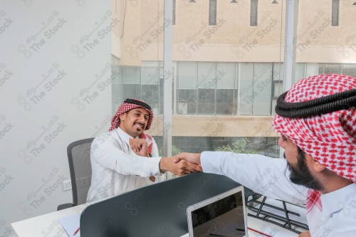 رجلان سعوديان يرتديان الزي السعودي التقليدي يتصافحان أثناء العمل في أروقة المكتب