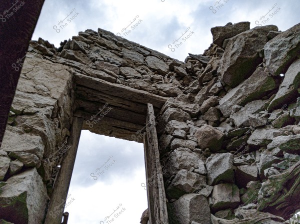 The image depicts the ruins of an old stone building, with walls made of large, unevenly placed stones. The cloudy sky is visible through large openings in the walls, and a stone door frame is seen without a door. The colors are somewhat dark with natural lighting.