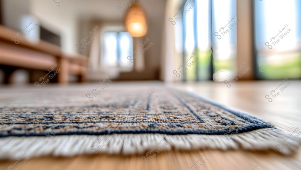 An image showing part of a modern living room with a focus on a carpet in beige and blue tones, featuring a simple pattern. The background is blurred, revealing large glass windows, warm hanging lighting from the ceiling, and wooden furniture.