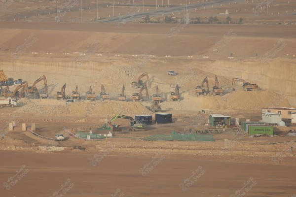 The image depicts a large construction site featuring a series of large excavators operating in a wide open excavation area. In the foreground, there are several mobile units and shipping containers, with a white truck on a road passing through the site. In the background, open desert landscapes and roads with a few vehicles are visible.