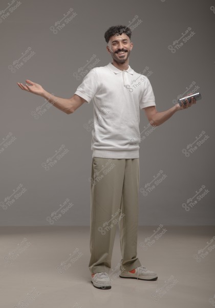 A man is standing and smiling, wearing a white polo shirt and loose beige pants, with white sneakers featuring orange stripes. He is holding a mobile phone in his left hand and his arms are extended open. The background is simple and dark gray in color.