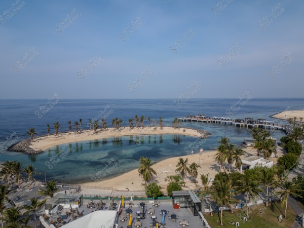 An aerial view of a beautiful beach overlooking a clear blue sky and the sea. The image shows a sandy beach shaped like a small peninsula adorned with palm trees and surrounded by sea water. There is a small pier extending over the water with structures on it. The area features a walkway with tables and chairs, with an atmosphere that feels tranquil and relaxing.