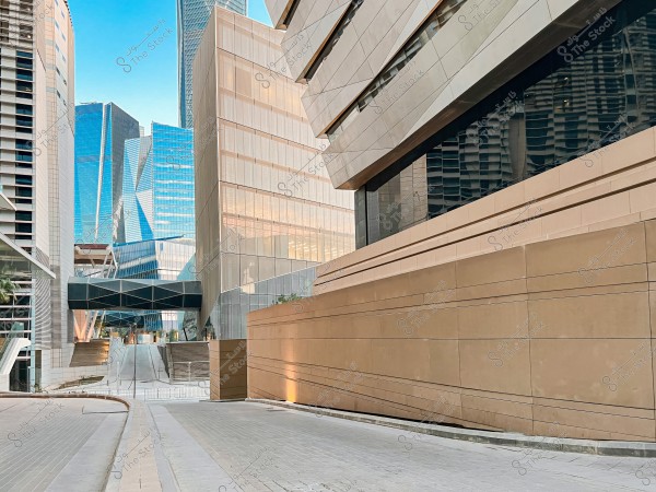 An image of a modern urban scene featuring a collection of glass and metal buildings with sleek, geometric designs. The scene includes a wide paved pathway flanked by buildings that reflect the clear blue sky. Elements of modern architecture and industrial materials like steel and glass are prominent.