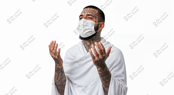 An image of a man wearing a white Ihram garment and a white medical face mask. His body and hands are covered with colorful tattoos. He is raising his hands as if in prayer. The background is white and clean.