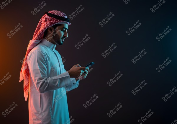 A portrait of a man wearing a traditional white thobe and a red and white checkered ghutra with a black agal, standing sideways while looking at his mobile phone. The background is dark with bright colors illuminating the image from the sides.