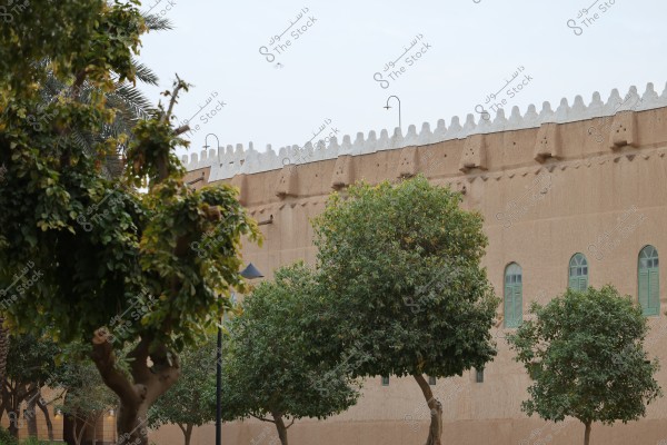 The image shows a section of a traditional building with tall turrets and distinctive architectural arches in the background. In the foreground, there are lush green trees with dense branches and some palm trees. The sky is clear with some lamps mounted on poles among the trees.