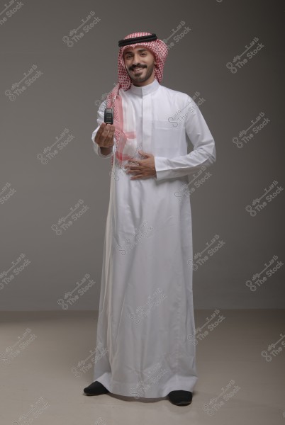 A portrait of a man standing and smiling, wearing traditional Saudi attire including a thobe, ghutra, and agal. He is holding a car key in his right hand and appears joyful. The background is a simple dark gray.