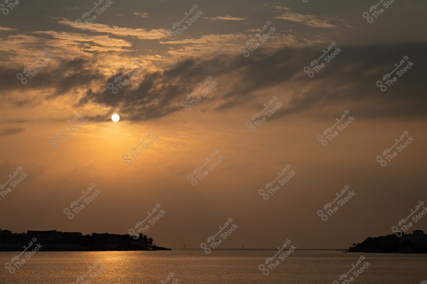 Image of a sunset over the sea with light reflections on the water. The sky is filled with shades of orange and gold with some clouds present. In the horizon, there is a silhouette of trees and buildings on both sides of the image.