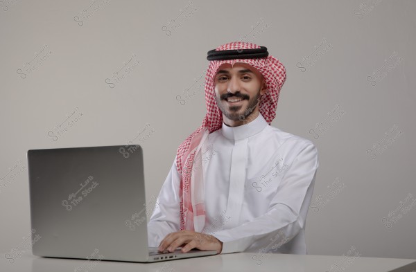 The image shows a man wearing traditional Saudi attire with a white thobe and red ghutra with a black agal, sitting in front of a laptop on a desk. The man is smiling and looking directly at the camera, with a light gray background.