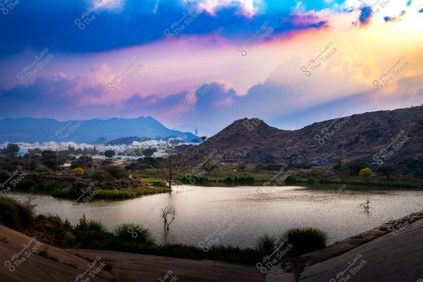 A scenic landscape featuring a water pond surrounded by grasses and trees, with a mountainous background. On the horizon, a cluster of white buildings is visible among rocky hills. The sky is covered with clouds colored in vibrant shades of blue and orange during sunset.
