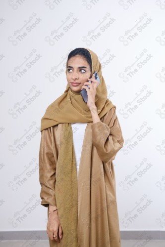 Portrait of a Saudi girl wearing an abaya on a white background holding a phone and making gestures with her hands while smiling, souvenir photos, documenting a happy moment.