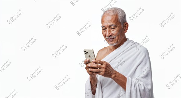 The image shows an elderly man with light skin and gray hair, wearing a white cloth resembling an ihram, looking at a smartphone in his hand. The background is white, highlighting the man\'s features and traditional clothing.