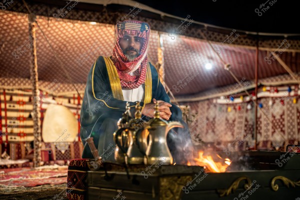 Image of a man sitting in a traditional tent illuminated with colorful lights in a nighttime setting. He is wearing traditional attire consisting of a white thobe, a black cloak, and a headscarf with red and white patterns. In the foreground, there are Arabic coffee dallahs on a lit fire. The ambiance suggests a traditional gathering in the Arabian Gulf region.