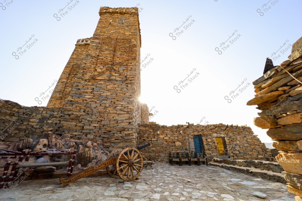 View of a traditional stone tower in an old village made from warm-toned earthy stones. An old wooden cannon with a wooden wheel is also visible, alongside a set of ornate wooden chairs. The blue and yellow doors enhance the earthy tones of the scene. The lighting reflects the clear daytime sun, casting shadows on the stone structure.